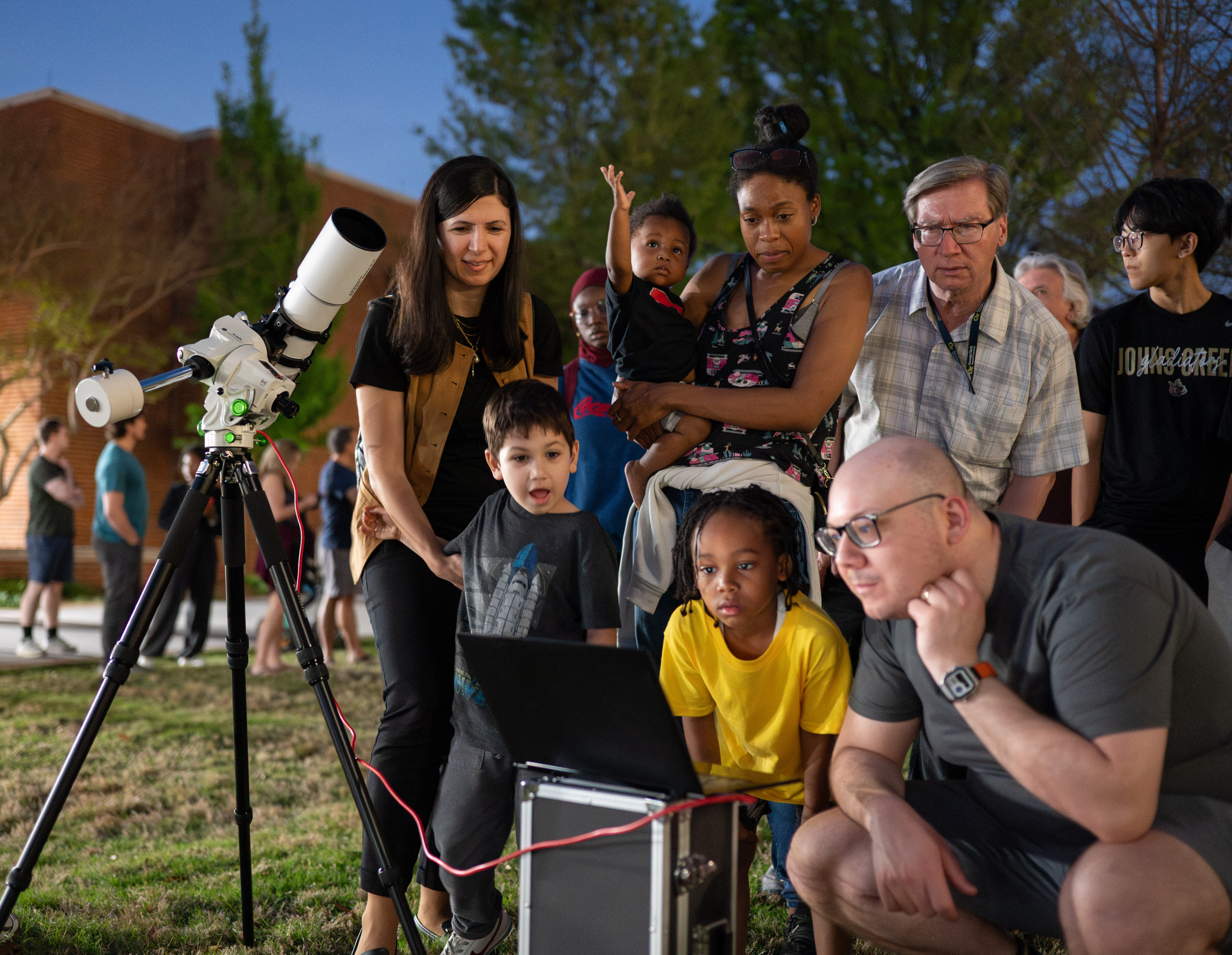 Attendees gather around a telescope and laptop during a public observatory night event held outdoors at dusk. Several adults and children stand in a group on a grassy area, watching images or data displayed on the laptop while a telescope on a tripod is positioned nearby. Trees and a brick building are visible in the background as other visitors mingle around the event.