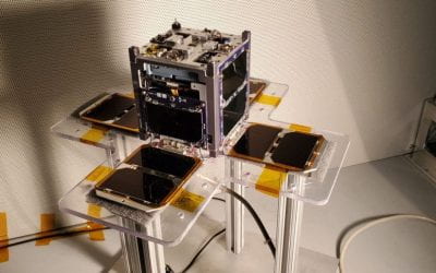 Small cube-shaped satellite prototype mounted on a clear stand, surrounded by several flat solar panel arrays arranged in a cross-like layout.