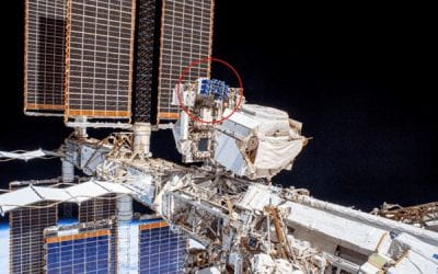 Exterior view of the International Space Station with large solar panels, highlighting a small experiment package mounted on the structure, circled in red.